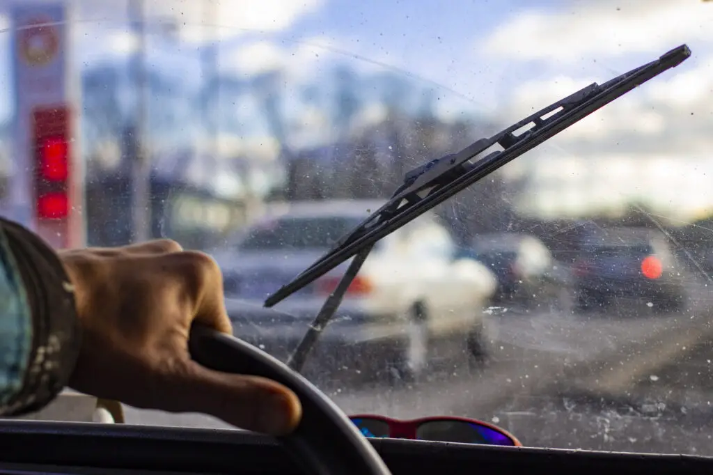 Person is driving a car with windshield covered with scratches and dirt. View from inside the car.